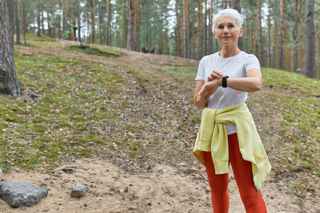 Outdoor portrait of confident active middle aged female in sportswear using smart watch monitoring pulse or heart rate while exercising in park.