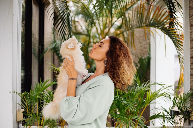 Free photo outdoor portrait of curly european tanned woman holds happy pet dog pomeranian spitz