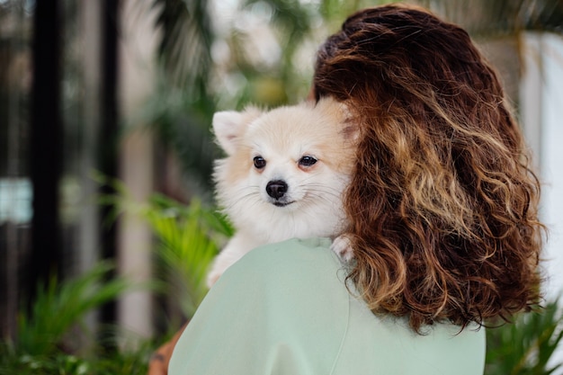 Outdoor portrait of curly european tanned woman holds happy pet dog pomeranian spitz