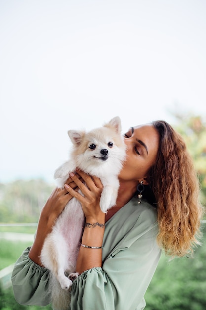 Outdoor portrait of curly european tanned woman holds happy pet dog pomeranian spitz