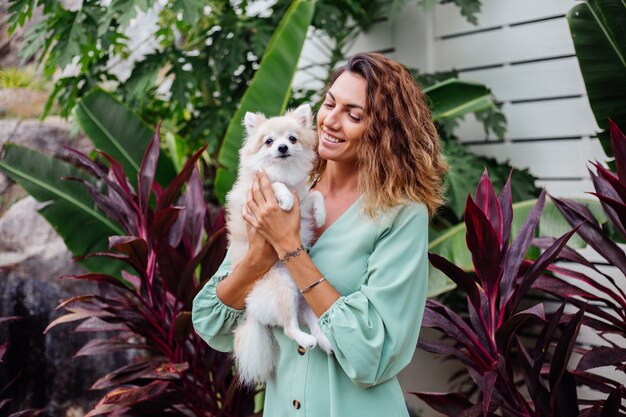 Outdoor portrait of curly european tanned woman holds happy pet dog pomeranian spitz