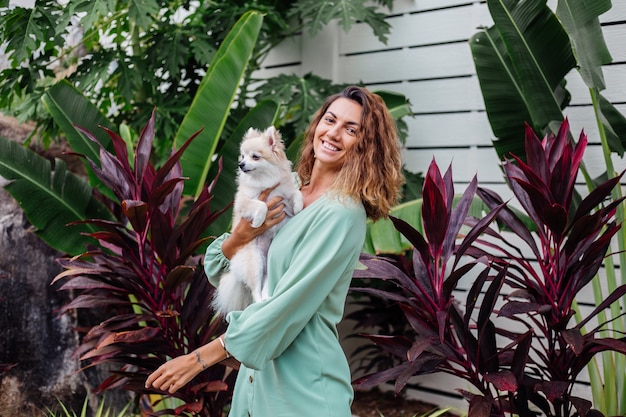 Outdoor portrait of curly european tanned woman holds happy pet dog pomeranian spitz