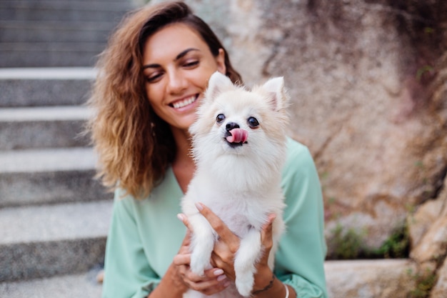 Outdoor portrait of curly european tanned woman holds happy pet dog pomeranian spitz