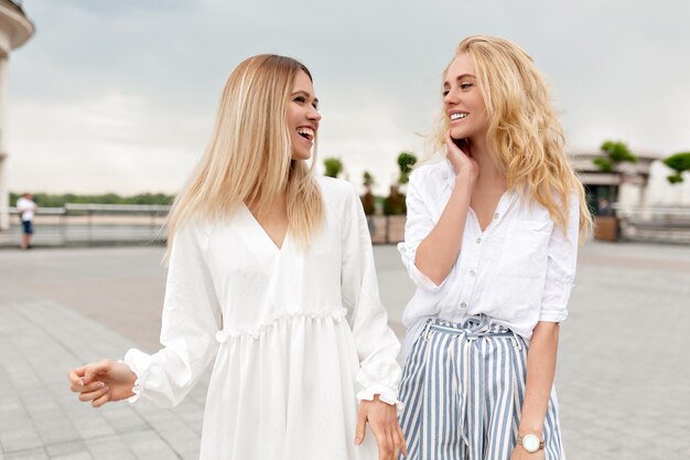 Outdoor portrait of cute ladies having fun outside relaxing in summer city while walking on the street and laughing on cloudy summer day. Two stylish girls outside