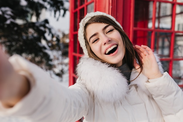 Free Photo outdoor portrait of cute woman with happy smile making selfie in london during winter vacation. adorable woman in white hat taking photo of herslef beside red phone booth..