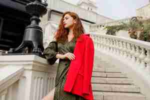 Free photo outdoor portrait of elegant ginger woman in green dress and red jacket standing on stairs near bridge