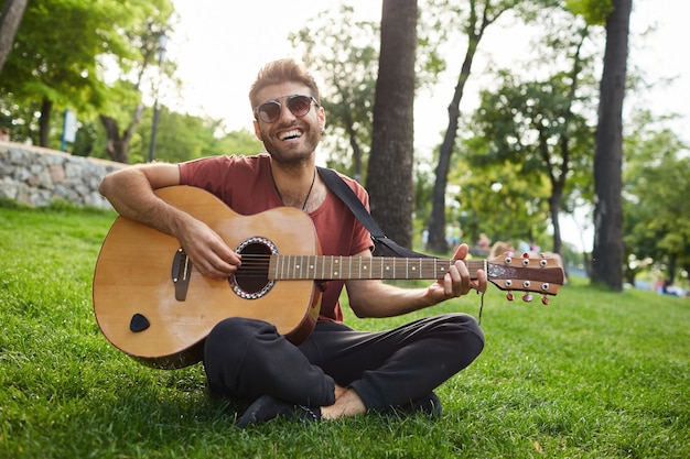 Outdoor portrait of handsome hipster guy sitting on grass in park and playing guitar