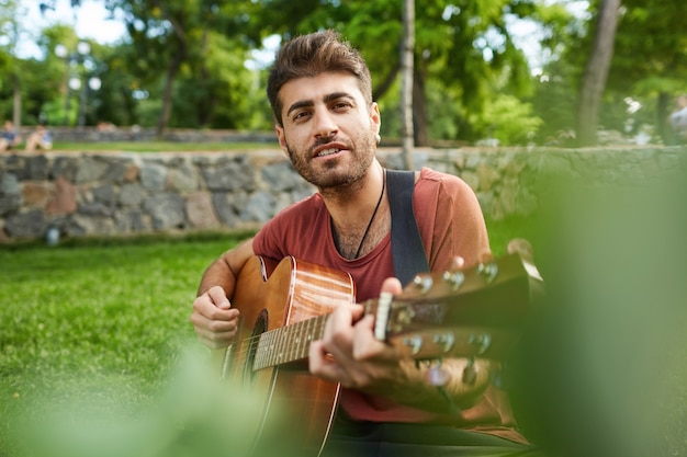 Outdoor portrait of handsome romantic guy sitting on grass in park and playing guitar