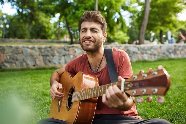 Outdoor portrait of handsome romantic guy sitting on grass in park and playing guitar