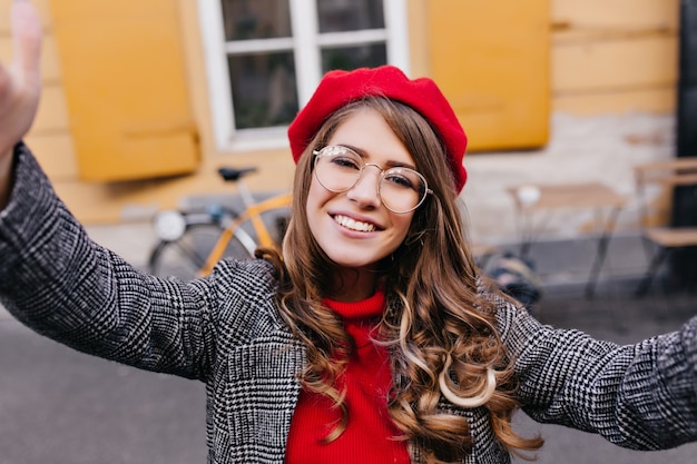 Free photo outdoor portrait of laughing romantic lady in glasses making selfie in front of yellow building