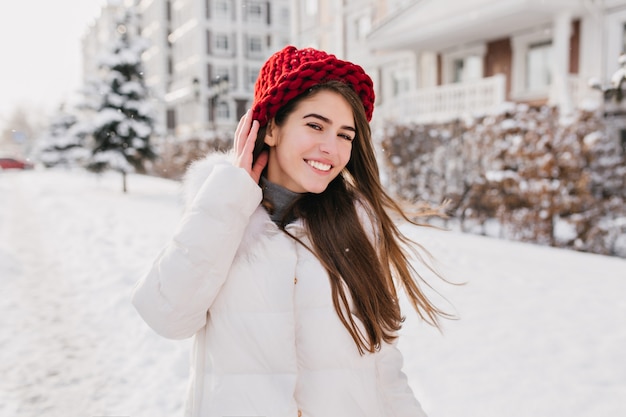 Outdoor portrait of pleased long-haired lady in red knitted hat walking down the street in snowy weekend. Photo of laughing cute lady in white winter coat having fun in cold morning..