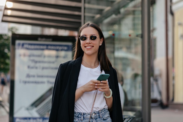 Outdoor portrait of pretty stylish girl in dark jacket in black glasses is scrolling smartphone and smiling while walking in the city in sunny warm day