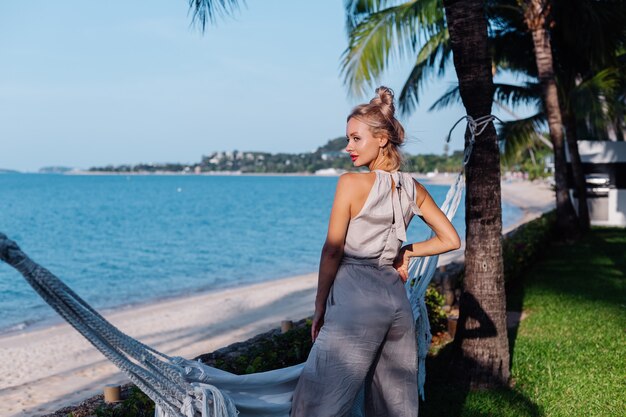 Outdoor portrait of rich caucasian woman in classic jumpsuit with red lipstick by hammock on vacation outside villa hotel