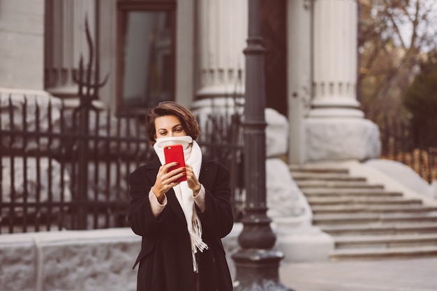 Free photo outdoor portrait of woman in black winter coat and white scarf in the street, holding mobile phone.