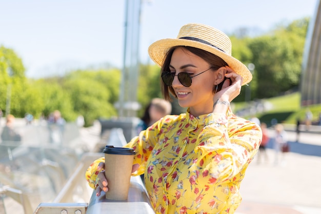 Outdoor portrait of woman in yellow summer dress and hat with cup of coffee enjoying sun, stands on bridge with city amazing view