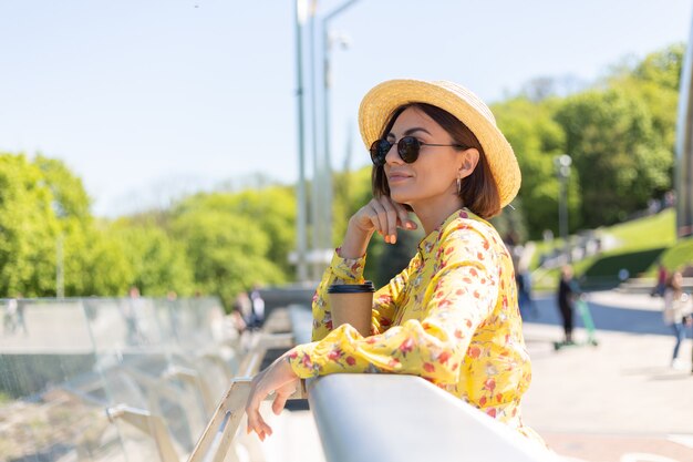 Outdoor portrait of woman in yellow summer dress and hat with cup of coffee enjoying sun, stands on bridge with city amazing view