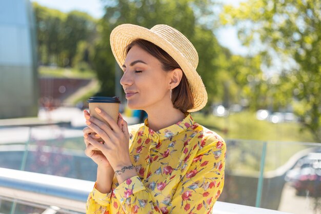 Free Photo outdoor portrait of woman in yellow summer dress and hat with cup of coffee enjoying sun