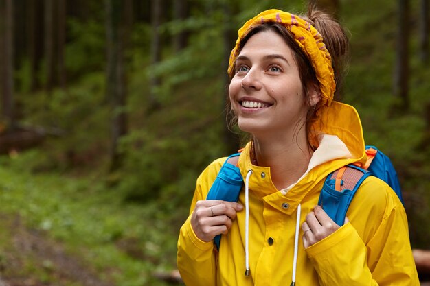 Outdoor shot of cheerful young woman looks thoughtfully into distance, wears yellow headband and raincoat, wanders in forest