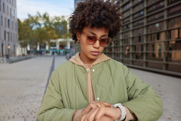 Free photo outdoor shot of curly haired woman wears sunglasses jacket and hoodie checks time on smartwatch spends free time walking in city during sunny weather uses application modern lifestyle concept