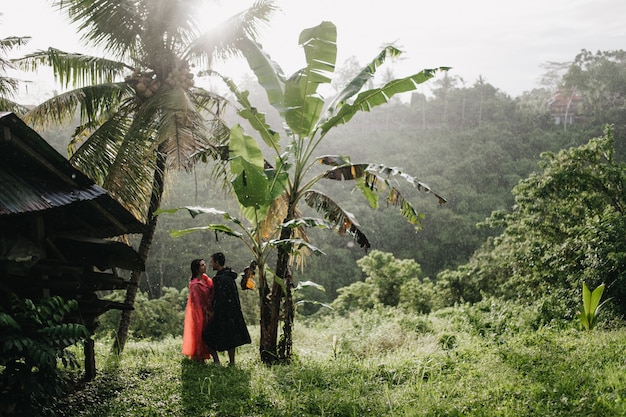 Outdoor shot of female tourist in pink raincoat posing with boyfriend on jungle. Portrait of couple chilling in tropical forest.