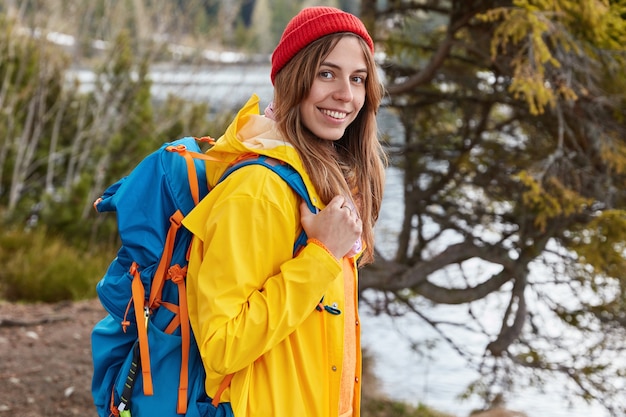 Free Photo outdoor shot of glad female with broad smile, stands sideways at camera, carries rucksack