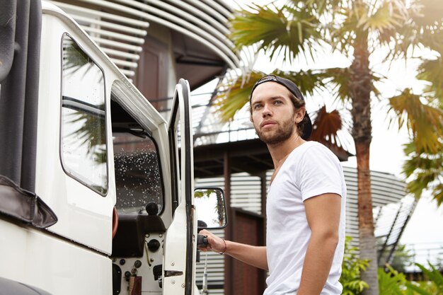 Outdoor shot of handsome young bearded man in T-shirt standing outside his white crossover utility vehicle, pulling its door handle and looking away, ready to get in