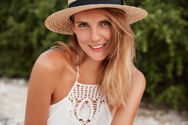 Outdoor shot of happy delighted female with pleasant look, wears summer white dress and hat, being in good mood. People, emotions and rest concept.