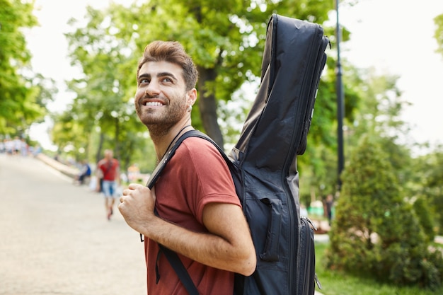 Outdoor shot of happy handsome hipster guy admire beautiful green park, walking with guitar, musician going on rehearsal