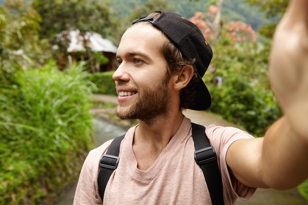 Free Photo outdoor shot of happy young hipster wearing backpack and baseball cap taking self-portrait, smiling and looking away. handsome traveler walking along country road