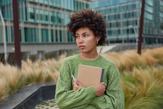 Outdoor shot of pensive curly haired young beautiful woman looks away dressed in casual green jumper holds notebook and pencil focused somewhere stands near modern urban building background