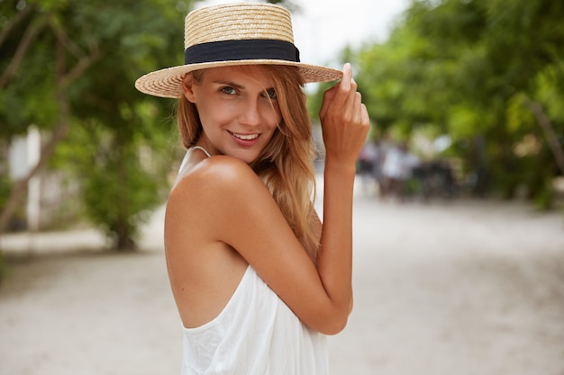 Outdoor shot of pleasant looking female with tanned healthy skin, dressed in white dress and summer hat, poses in park with confident satisfied expression