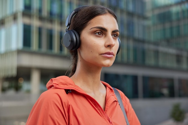 Free photo outdoor shot of serious brunette young woman leads sporty lifestyle listens music via headphones enjoys playlist exercises in city dressed in anorak poses at street takes break after workout