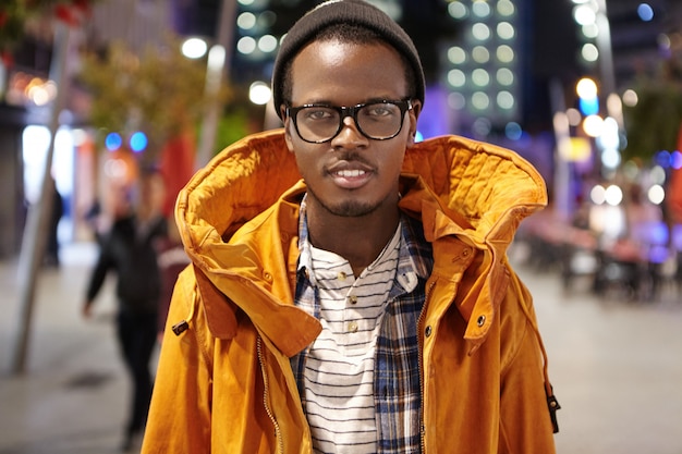 Outdoor shot of stylish dark-skinned tourist in winter coat, hat and glasses walking around night city, hitchhiking across Europe, standing in the middle of street and looking with smile