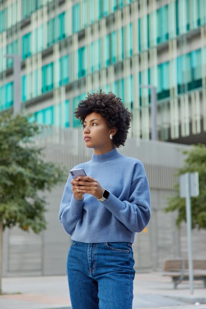 Outdoor shot of thoughtful beautiful woman wears casual blue jumper and jeans stands against urban setting uses mobile phone connected to wireless internet going to call taxi strolls in city