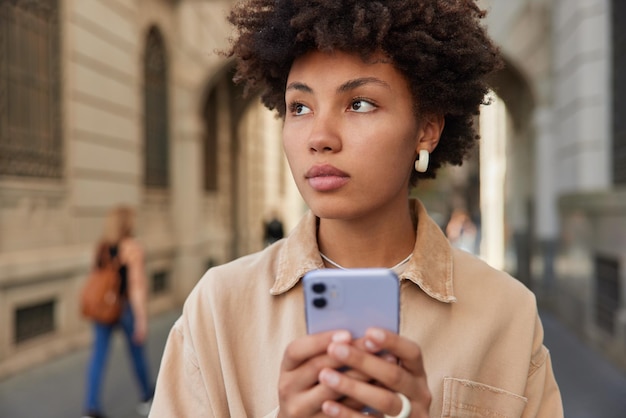 Outdoor shot of thoughtful curly haired beautiful woman browses mobile phone uses navigation app walks on city street during travel journey sends content shares multimedia online focused away