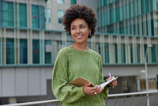 Outdoor shot of thoughtful curly haired woman stands against urban building writes information in notebook holds pen and notepad notes text creats new chapter o book dressed in casual green jumper