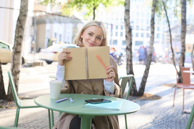 Free photo outdoor shot of young beautiful blond girl with notebook woman sitting in outdoor cafe writing in