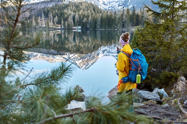 Free Photo outdoor shot of young traveler with bag, stands back to camera, enjoys mountains, fresh air and small lake