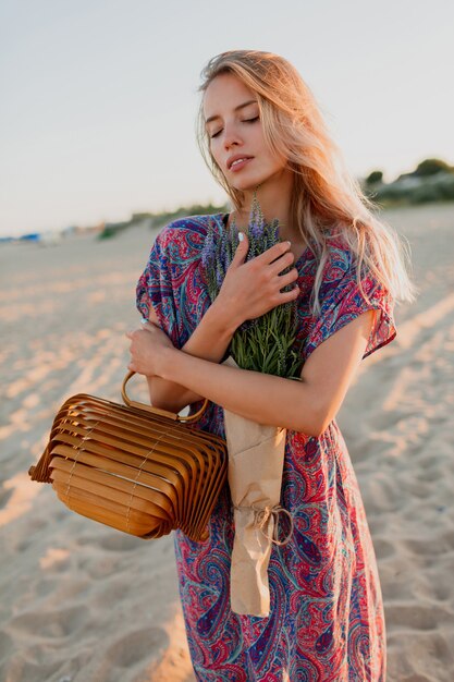 Outdoor summer image of beautiful romantic blond woman in colorful  dress walking on the beach with bouquet of lavender.