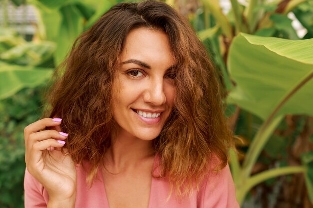 Outdoor summer portrait of seductive brunette woman with wavy hairs in pink dress posing on tropical