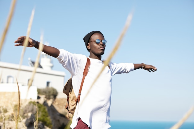 Outdoor summer shot of carefree happy young traveler standing at seaside with arms outstretched