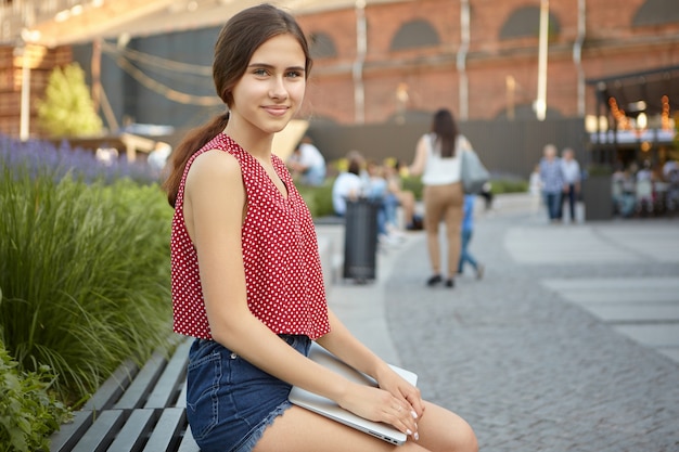 Outdoor summer view of beautiful positive young woman in dotted red top and jeans shorts using portable electronic device in park, looking and smiling . Modern technology and communication