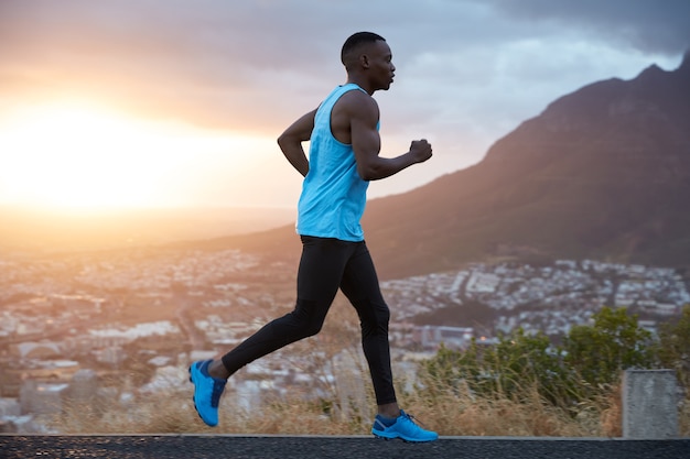 Outdoor view of active young male jogger covers long destiantion ealy in morning at dawn, runs over mountains view, has biceps, dressed in activewear, breathes deeply, enjoys summer weather.