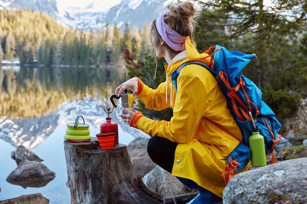 Free photo outdoor view of young woman uses tourist equipment for making coffee, has portable gas stove on stump