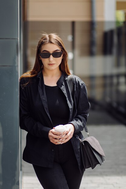 Outdoors lifestyle portrait of stunning brunette girl. Drinking coffee and walking on the city street.