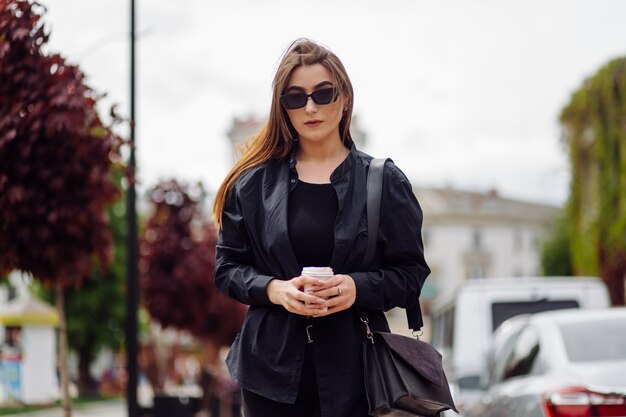 Outdoors lifestyle portrait of stunning brunette girl. Drinking coffee and walking on the city street.