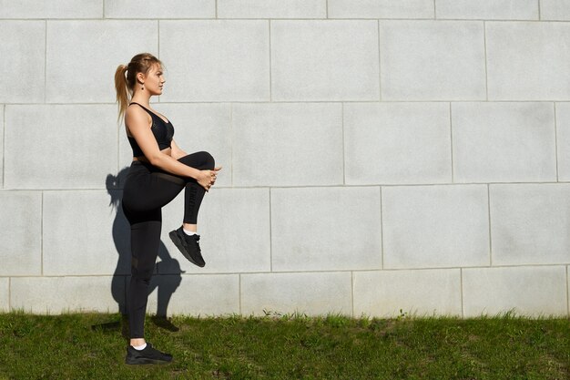 Outdoors summertime portrait of attractive young female in sportswear stretching quadricep, standing on grass against blank wall background with copy space for your text or advertising content