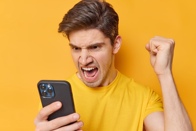 Free photo outraged angry adult man screams loudly focused at smartphone screen clenches fist has argument with something during distance call dressed in casual t shirt isolated over yellow background