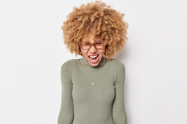 Outraged emotional young curly woman screams out angrily expresses rage hates something quarrel with someone wears turtleneck and spectacles isolated over white background. Negative emotions