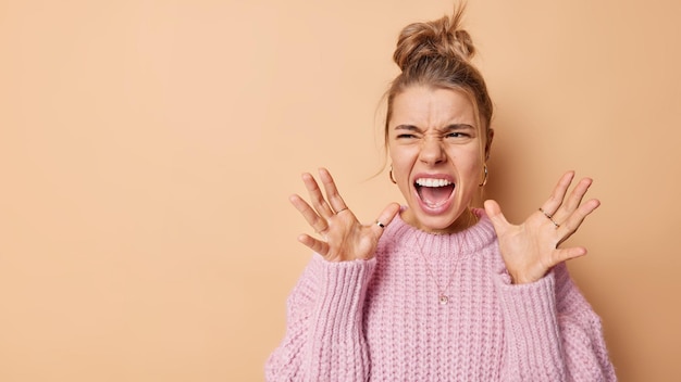 Outraged European woman screams from anger raises palms with irritation expresses negative emotions wears knitted sweater isolated over beige background blank space on left to place your text